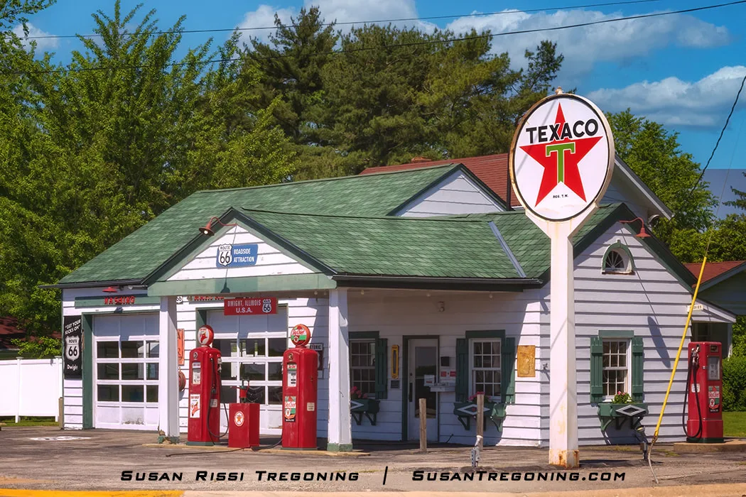 A vintage Texaco gas station with a white exterior and green roof stands along Route 66 in Dwight, Illinois. Two classic red gas pumps sit in front of the building, and a tall Texaco star sign rises beside them. Signs on the building identify it as a Route 66 roadside attraction.