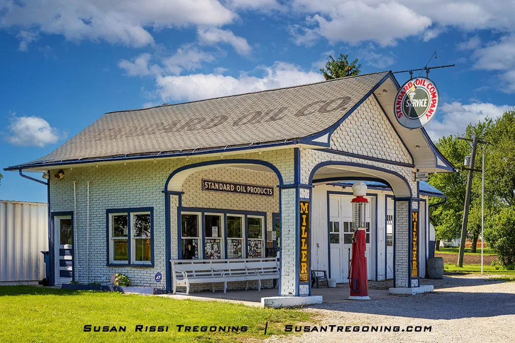 A light‑blue Standard Oil gas station with dark‑blue trim stands under a partly cloudy sky. A round Standard Oil of Ohio Service sign hangs from the roofline, and a red vintage gas pump sits near the entrance. Yellow vertical lettering reading Miller Tires appears on the front corner pillars, and a white bench rests beneath the front windows. Trees and grass surround the building.