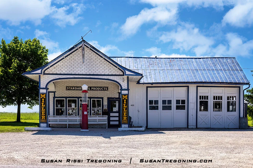 A white Standard Oil gas station with blue trim stands under a bright sky. A single vintage red gas pump sits beneath the covered entrance, and vertical Miller Tires signs appear on both sides of the doorway. Two garage doors are visible on the right side of the building. The station now serves as the Odell Route 66 Welcome Center.
