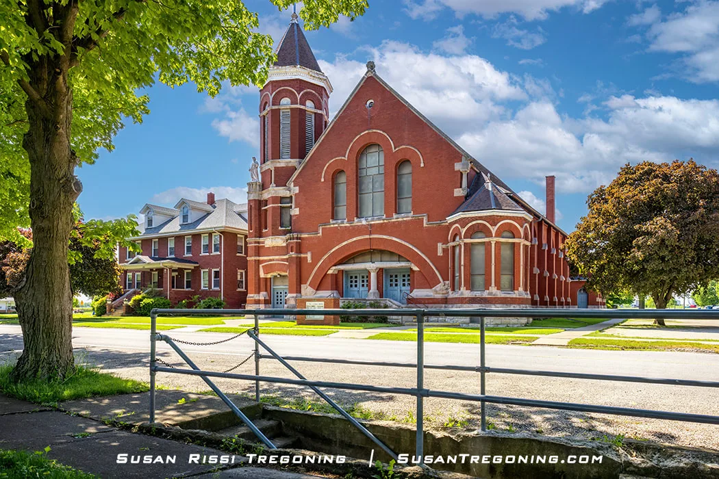 A large red‑brick church with Romanesque architectural features stands along a sunny street in Odell, Illinois. The building has arched windows, decorative stone trim, and a tall corner tower with a conical roof. Trees, a sidewalk, and a neighboring house appear nearby. The church is located across the street from the historic Pedestrian Subway Tunnel on Route 66.