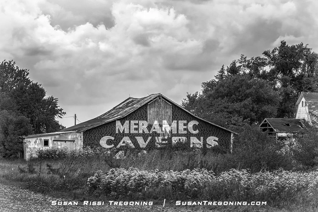 A weathered barn with large painted lettering reading Meramec Caverns stands amid overgrown vegetation. The structure has faded boards and a deteriorated roof. Dark, heavy storm clouds fill the sky above, and part of another aging building appears to the right.