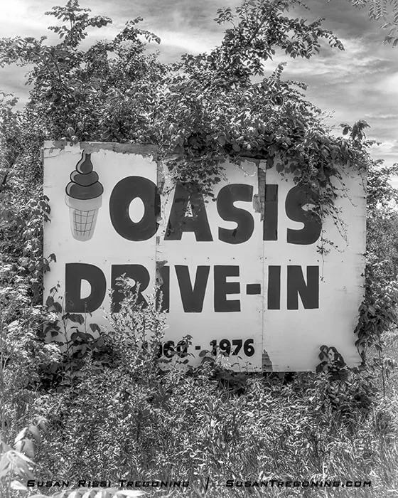 A weathered billboard reading Oasis Drive‑In stands amid tall grass and dense overgrown vegetation. The faded sign includes an illustration of an ice‑cream cone and the dates 1961–1976. The black‑and‑white image shows the sign partially obscured by vines and surrounded by natural growth.