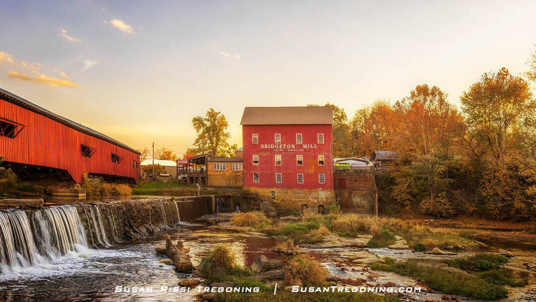 The red Bridgeton Mill stands beside Big Raccoon Creek near a small waterfall, with the Bridgeton Covered Bridge extending across the water to the left. Autumn trees surround the mill and bridge under a warm sunset sky.