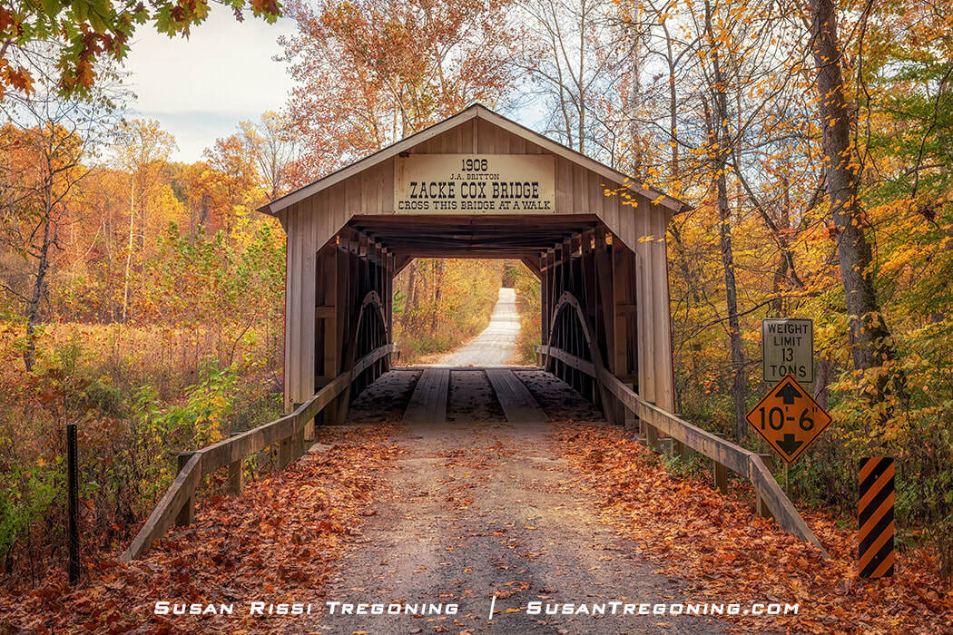 #20 Zacke Cox Covered Bridge, a red wooden bridge with white trim, surrounded by autumn foliage in shades of yellow and orange, with a gravel road and posted weight‑limit and clearance signs at the entrance.