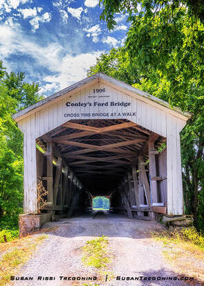 The Conley’s Ford Covered Bridge stands along a gravel road surrounded by green trees, with its white exterior and 1906 date board above the entrance. The bridge originally featured Daniels Arch portals, but these were replaced with Britton Portals during a 1991 renovation.
