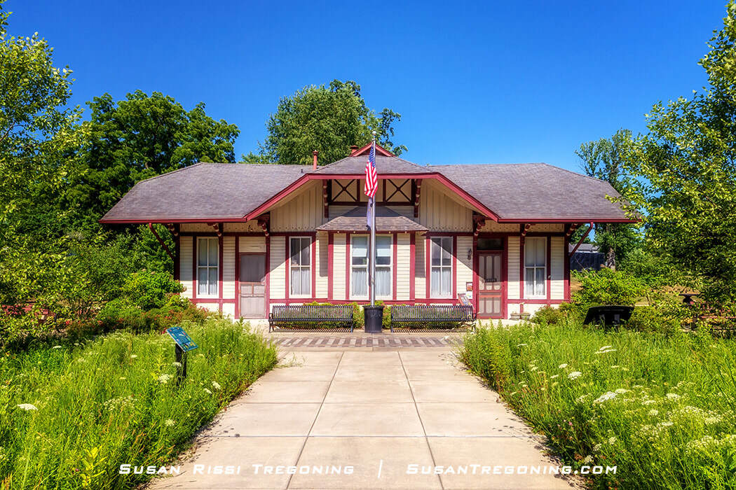 The former Rockville train depot, now the Parke County Visitor Center, is a small cream‑colored building with red trim, a dark roof, and a central entrance, set along a paved walkway with surrounding greenery and an American flag in front.