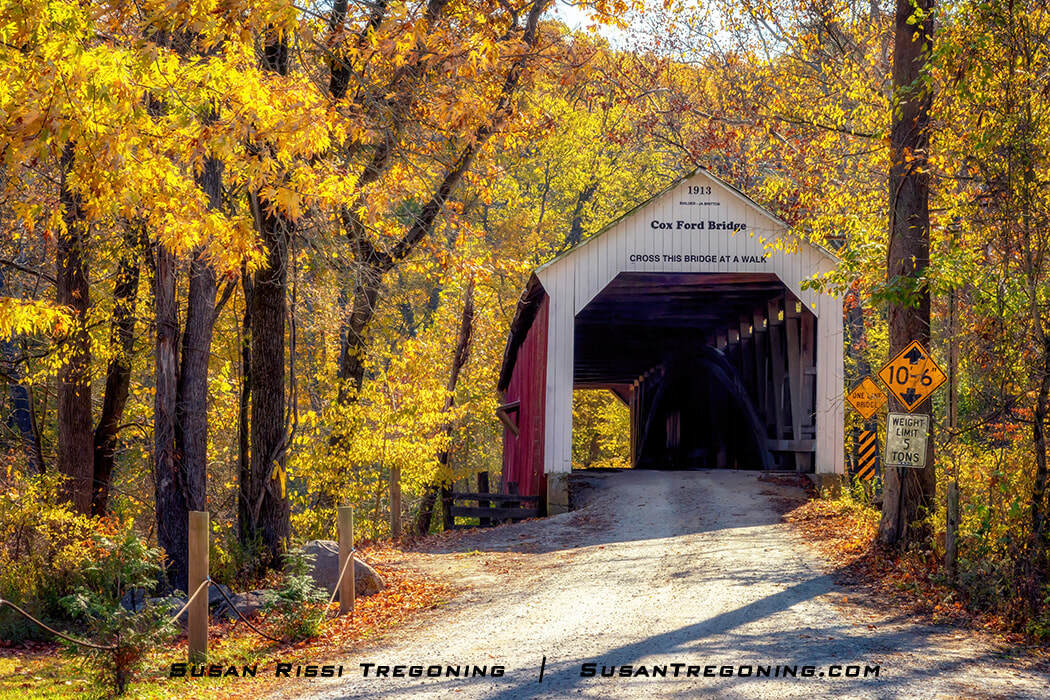 Cox Ford Covered Bridge, a white‑entrance, red‑sided wooden span dated 1913, set along a gravel road and framed by golden autumn foliage with posted clearance and weight‑limit signs at the entrance.