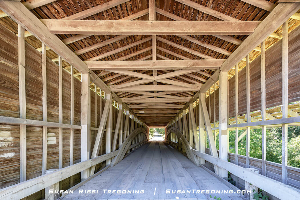 The interior of the Portland Mills Covered Bridge shows a wooden plank floor, vertical and diagonal support beams, and exposed trusses leading toward the bright opening at the far end, with greenery visible outside.
