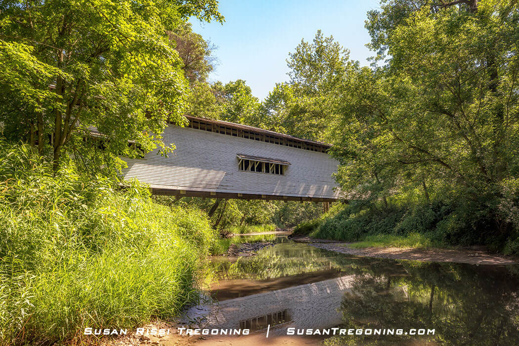 The white Portland Mills Covered Bridge, shown in profile, reflects in Little Raccoon Creek. It is the oldest Parke County, Indiana, Covered Bridge. 