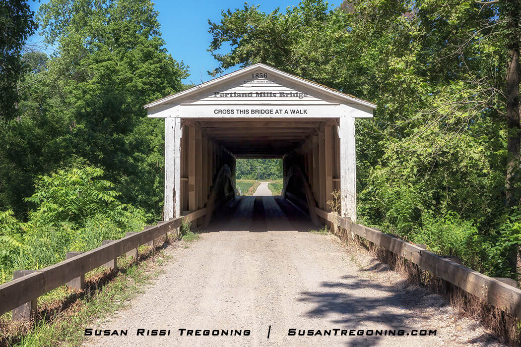 The white Portland Mills Covered Bridge stands among dense green trees, with its 1856 date board visible above the entrance. Some of the original bridge siding has been reused on the portals, showing older weathered boards alongside newer material.