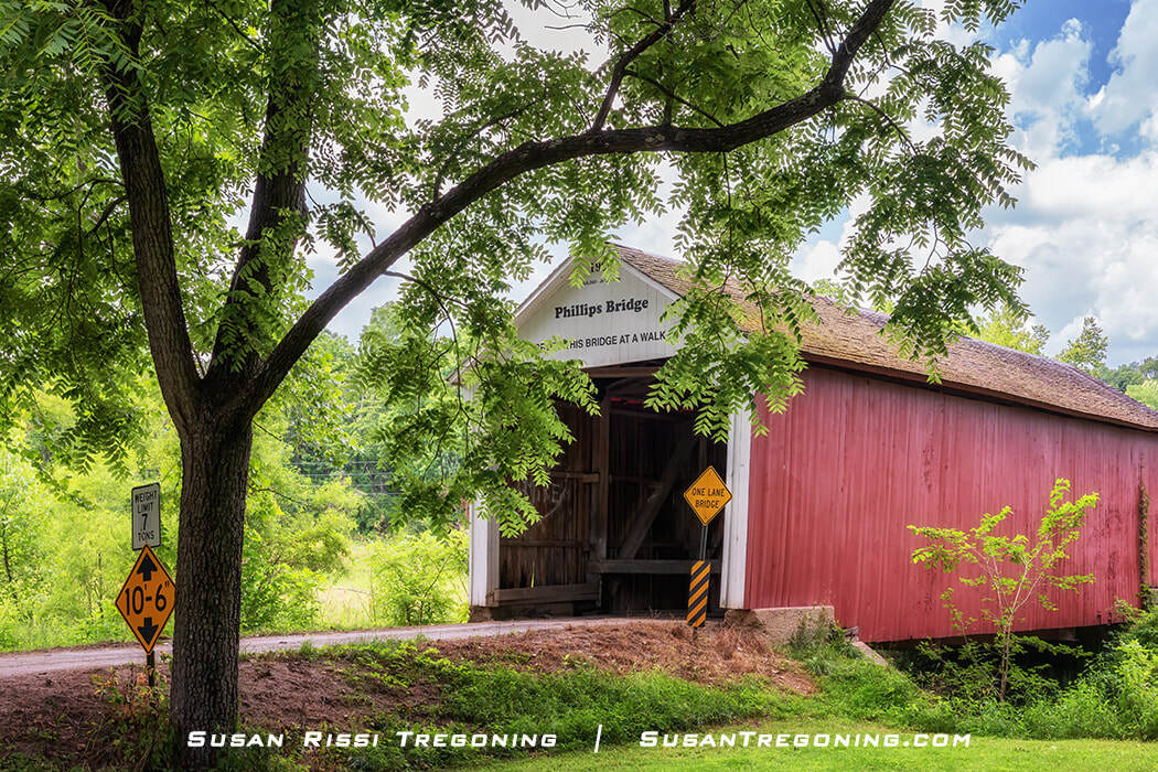   
#22 Phillips Covered Bridge, a red wooden bridge with a white gabled entrance, set along a dirt road and surrounded by lush green trees, with posted clearance and weight‑limit signs near the entrance.