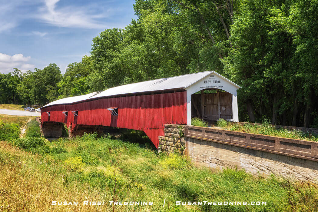 The south portal of the West Union Covered Bridge, a red wooden span with a white gabled entrance dated 1876, crossing Sugar Creek and surrounded by green trees under a clear sky.