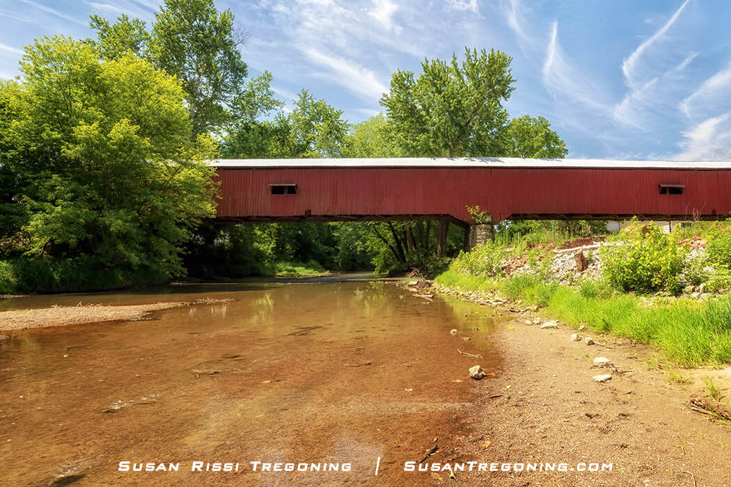 A profile view of the red Mansfield Covered Bridge spans Big Raccoon Creek, with its white roof and small side openings visible above the calm, shallow water. Green trees surround the bridge under a bright blue sky with wispy clouds.