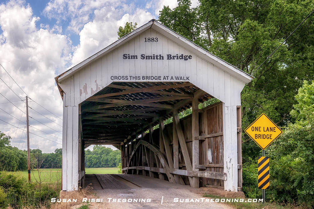 A white covered bridge labeled “Sim Smith Bridge” shows J. A. Britton’s pointed “Britton Portal” and one of his early Burr Arch trusses, with the arch coming to a sharp peak before he refined the design around 1900. A yellow “One Lane Bridge” sign stands beside the entrance, with trees and grass surrounding the bridge under a partly cloudy sky.