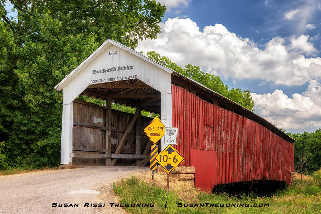 Sim Smith Covered Bridge, a red wooden bridge with a white gabled entrance dated 1883, surrounded by green trees and posted with one‑lane, weight‑limit, and clearance signs at the approach.