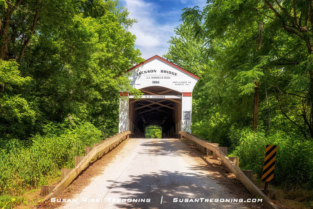 Jackson Covered Bridge, a long white‑and‑red covered bridge dated 1861, surrounded by green trees, with posted clearance and safety signs at the entrance.