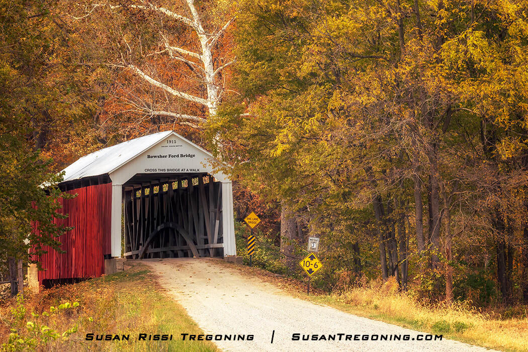 Bowsher Ford Covered Bridge, a red wooden span with white trim and a peaked roof, set along a gravel road and surrounded by yellow and orange autumn foliage with posted one‑lane and clearance signs at the entrance.