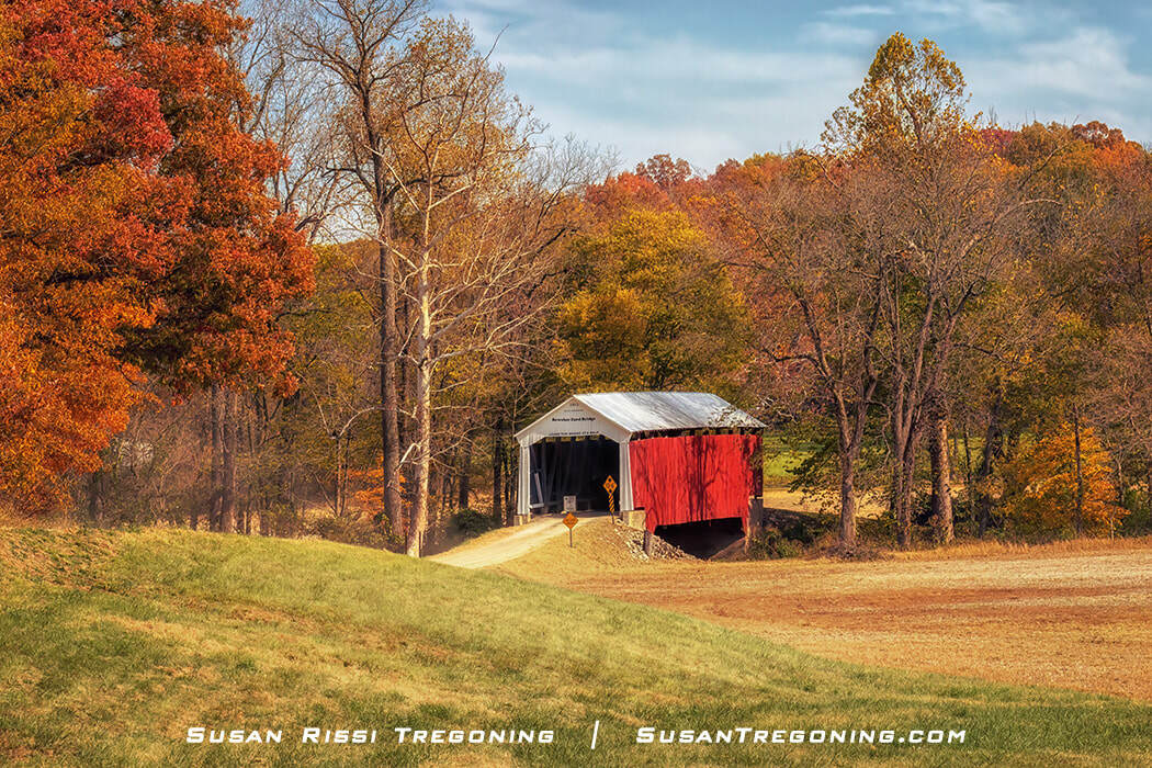 Bowsher Ford Covered Bridge seen from a distance, a red wooden span with a white roof set among open fields and autumn trees under a partly cloudy sky.