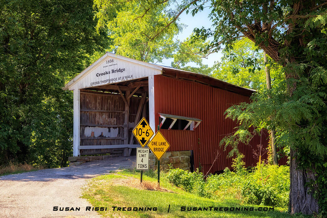 Red wooden Crooks Covered Bridge with white trim, built in 1856, spanning a creek and surrounded by green summer trees. Clearance, one‑lane, and weight‑limit road signs stand in front of the entrance.