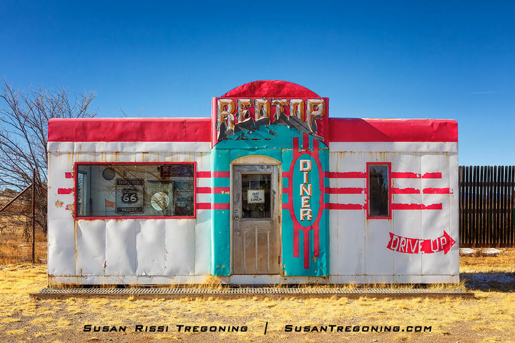 The Red Top Valentine Diner near Route 66 in Edgewood, New Mexico, shown as a small vintage diner with a red‑and‑white exterior, turquoise accents, and a “Red Top Diner” sign above the door. A painted arrow on the side reads “Drive Up,” and a sign on the door says “Out for Lunch,” all set against a clear blue sky.