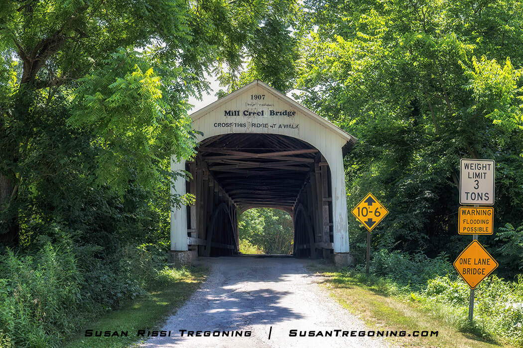   
Mill Creek Covered Bridge, a white wooden span with a peaked roof and posted clearance and weight‑limit signs, set along a tree‑lined road and surrounded by lush green summer foliage.