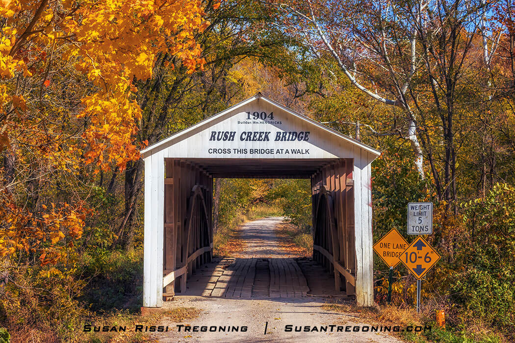 Rush Creek Covered Bridge, a white wooden span with a peaked roof set along a gravel road, surrounded by yellow and orange autumn foliage with posted weight‑limit and clearance signs at the entrance.