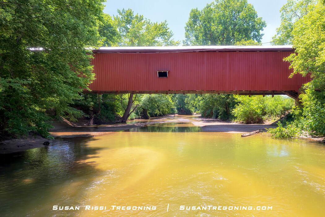 Crooks Covered Bridge shown in profile as it spans Little Raccoon Creek, surrounded by green summer trees and reflected in the calm water below.