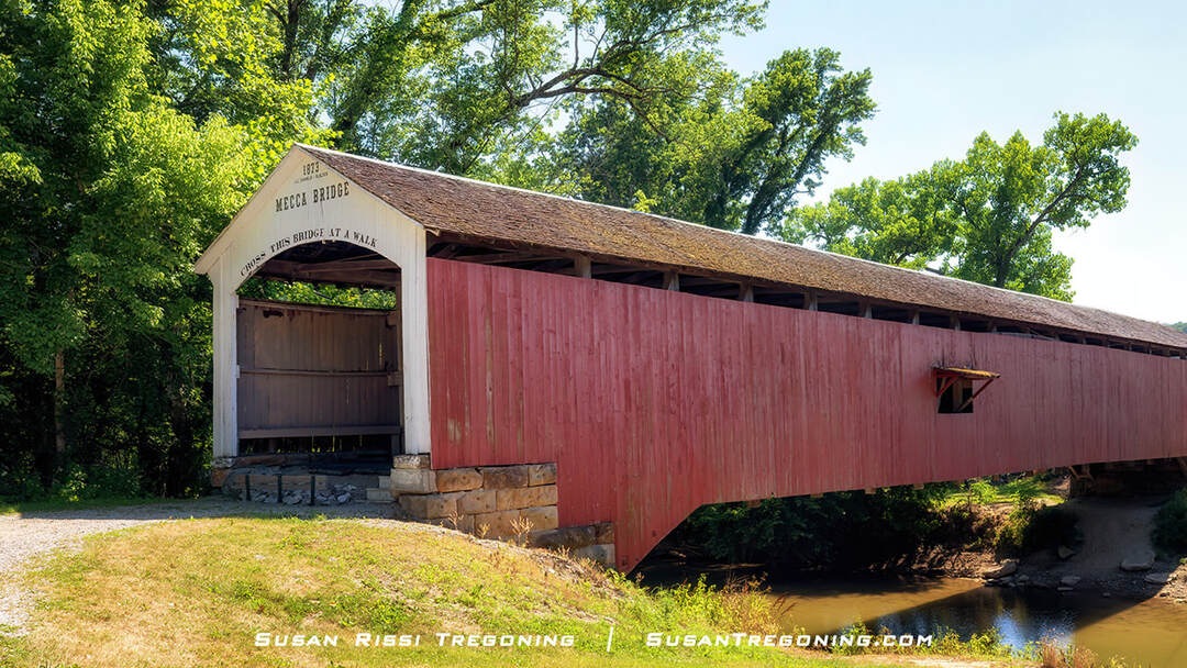 Mecca Covered Bridge, a red wooden bridge with a white gabled entrance dated 1873, spanning a small creek and surrounded by green trees, with a gravel path leading to the bridge.