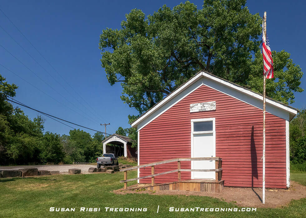 The Wabash Township Number 3 one‑room schoolhouse, a small red building with white trim and a front ramp, sits beside the Mecca Covered Bridge, with green trees, grass, and a clear blue sky in the background.