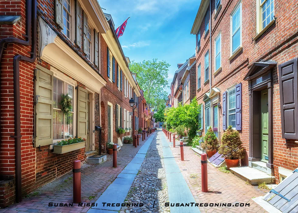 A view looking down the cobblestone street of Elfreths Alley in Philadelphia, showing a narrow lane lined with historic brick row homes. The perspective is from the far end of the street, with the buildings and walkway forming a calm, linear view toward the opposite end.