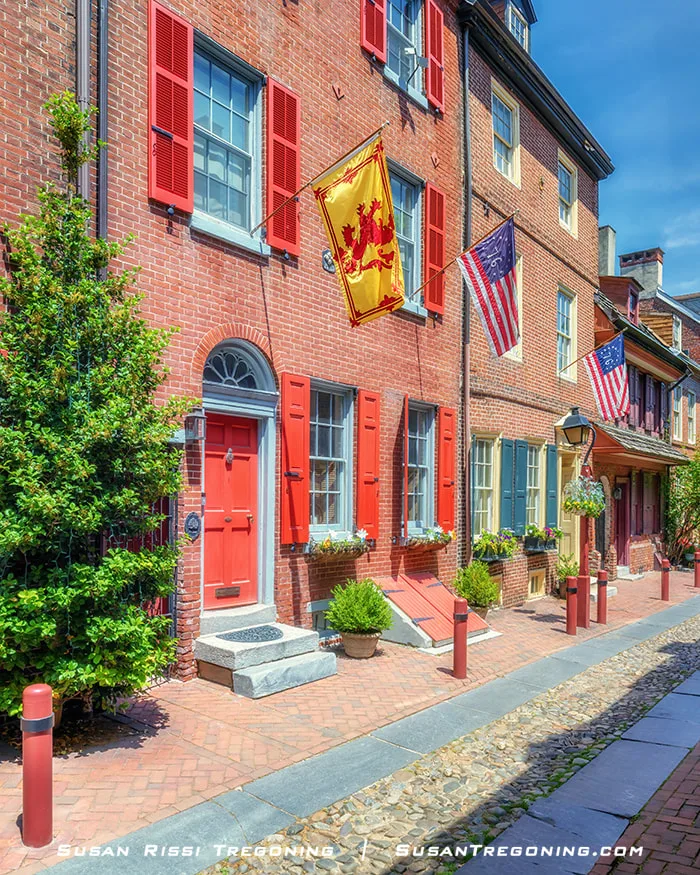A Colonial‑era street lined with brick buildings featuring assorted period flags, flower boxes, brightly colored doors and windows, and detailed brickwork along the facades.
