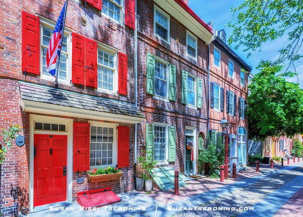 A narrow brick street lined with historic 18th‑century row homes in Elfreths Alley in Philadelphia. The homes have brick facades, shuttered windows, and doorways close to the sidewalk. The alley curves slightly into the distance, showing a quiet pedestrian walkway with no vehicles present.

