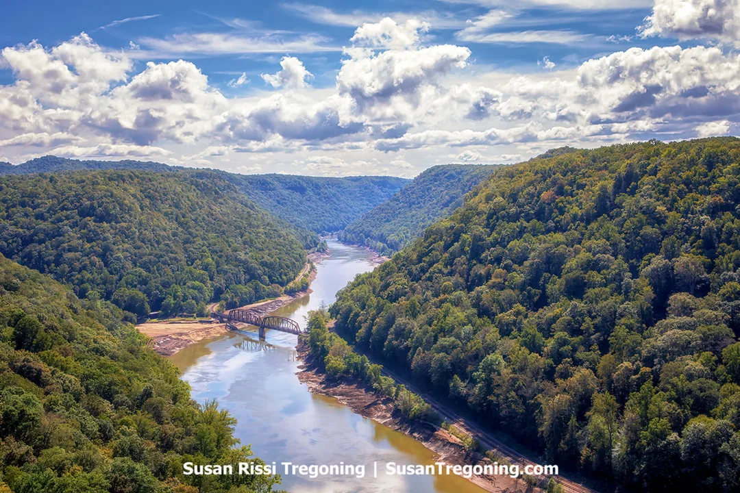 Overlooking the New River Gorge from Hawks Nest State Park in West Virginia, with a steel bridge spanning the river between steep, forested hillsides under a partly cloudy sky.