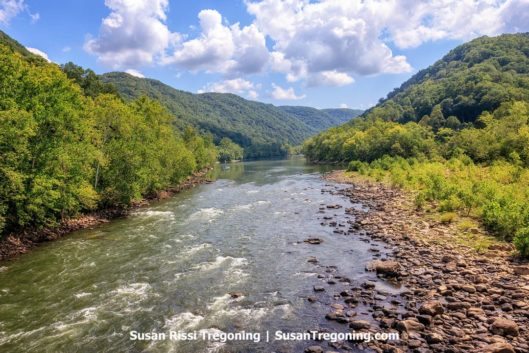 View from the Thurmond Railroad Bridge looking across the New River toward the surrounding Appalachian Mountains, with a small raft visible in the distance on the river below.