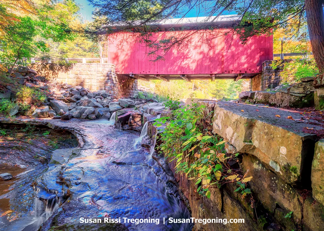 A red covered bridge with white trim spans a rocky creek and small waterfall, surrounded by early autumn foliage in shades of green, yellow, and orange. The bridge rests on stone abutments above cascading water, with large rocks and plants visible along the creek. The location is widely regarded as one of the most scenic covered bridge settings in Pennsylvania.