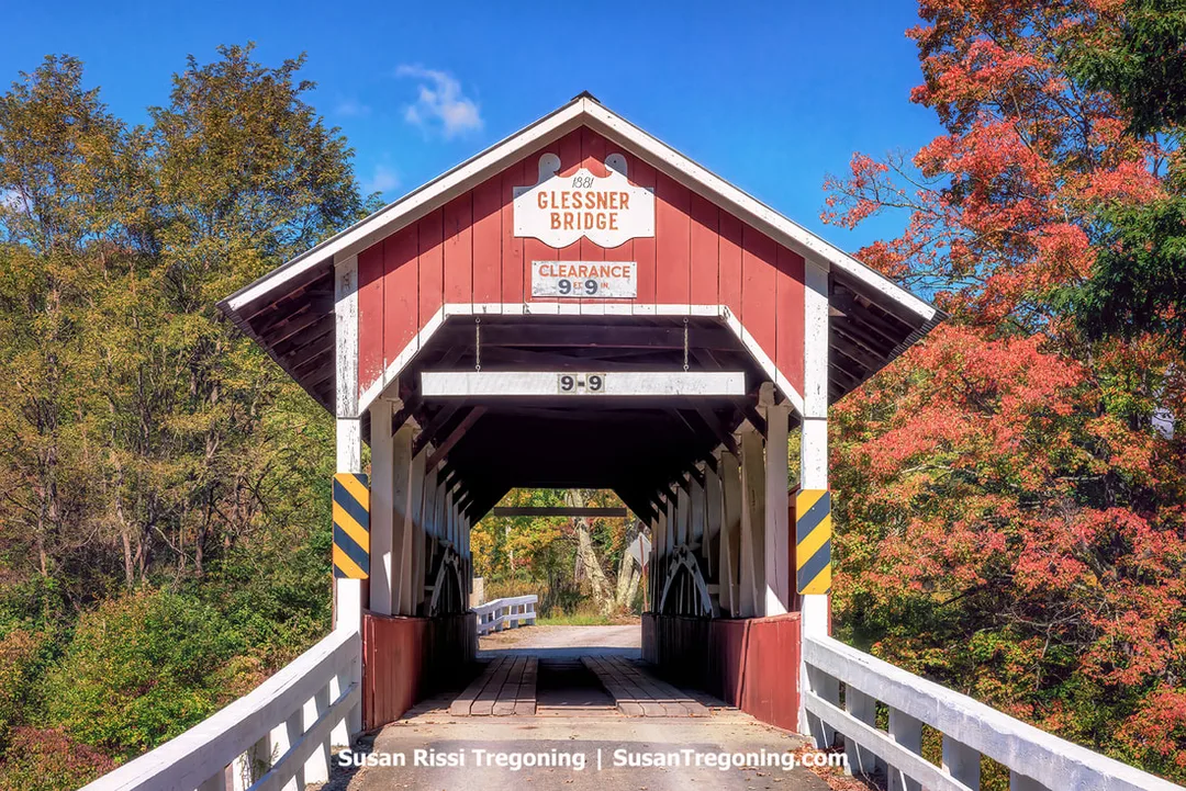 
A red covered bridge with white trim stands above a narrow creek, surrounded by autumn trees in shades of yellow, orange, and green. The bridge has a peaked roof and black‑and‑yellow caution stripes at the entrance. A sign above the portal reads 1881 Glessner Bridge with a posted clearance of 9 feet 9 inches.