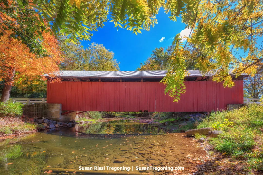 A red covered bridge with white trim spans a small creek, viewed from the side with its wooden structure and stone abutments visible. Autumn trees in shades of orange, yellow, and green surround the bridge under a blue sky. The structure is a replica and the only covered bridge in Somerset County not listed on the National Register of Historic Places.