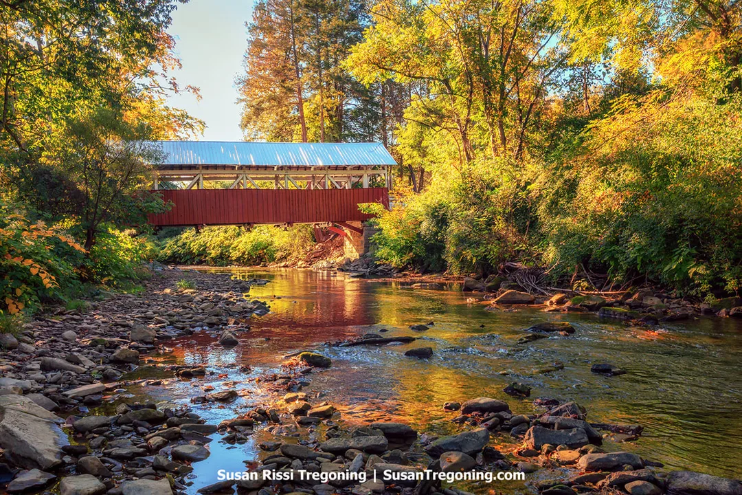 A red covered bridge with a metal roof spans a shallow rocky creek surrounded by trees with autumn foliage in shades of yellow, orange, and green. Sunlight filters through the leaves, creating warm reflections on the water and illuminating the wooden trusses beneath the bridge.