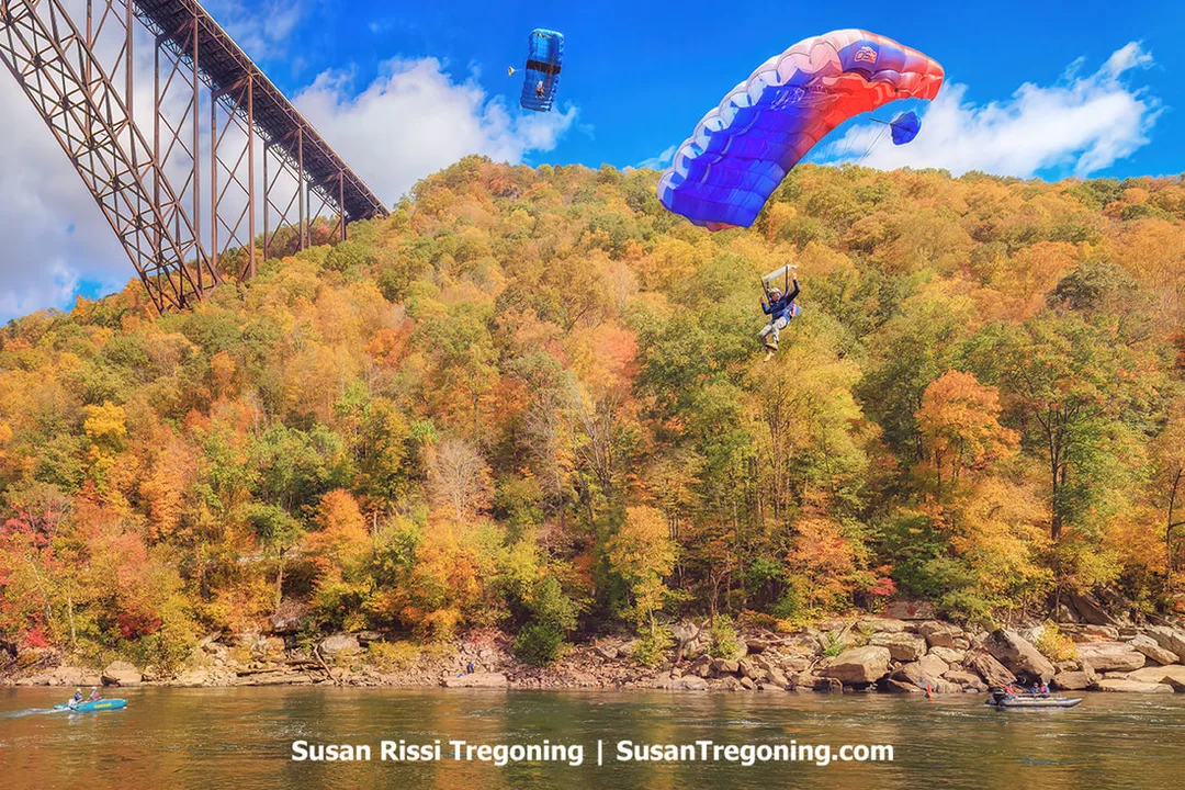 Two parachutists descend near a large steel bridge, one under a blue and red canopy and the other under a blue canopy, above a river and forested hillsides covered in autumn foliage beneath a bright blue sky with scattered clouds.