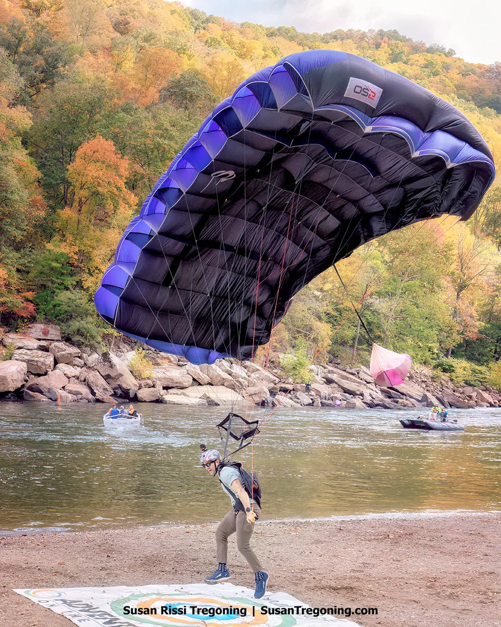 A BASE jumper descends toward a circular landing target beside a river, with his feet hovering a few feet above the marked bullseye as a dark purple canopy remains inflated overhead and autumn foliage fills the background.