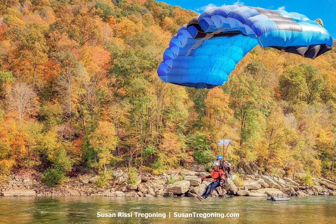 A tandem pair descends under a bright blue canopy toward a riverbank, with autumn‑colored hills rising behind them and a small boat visible on the water below.