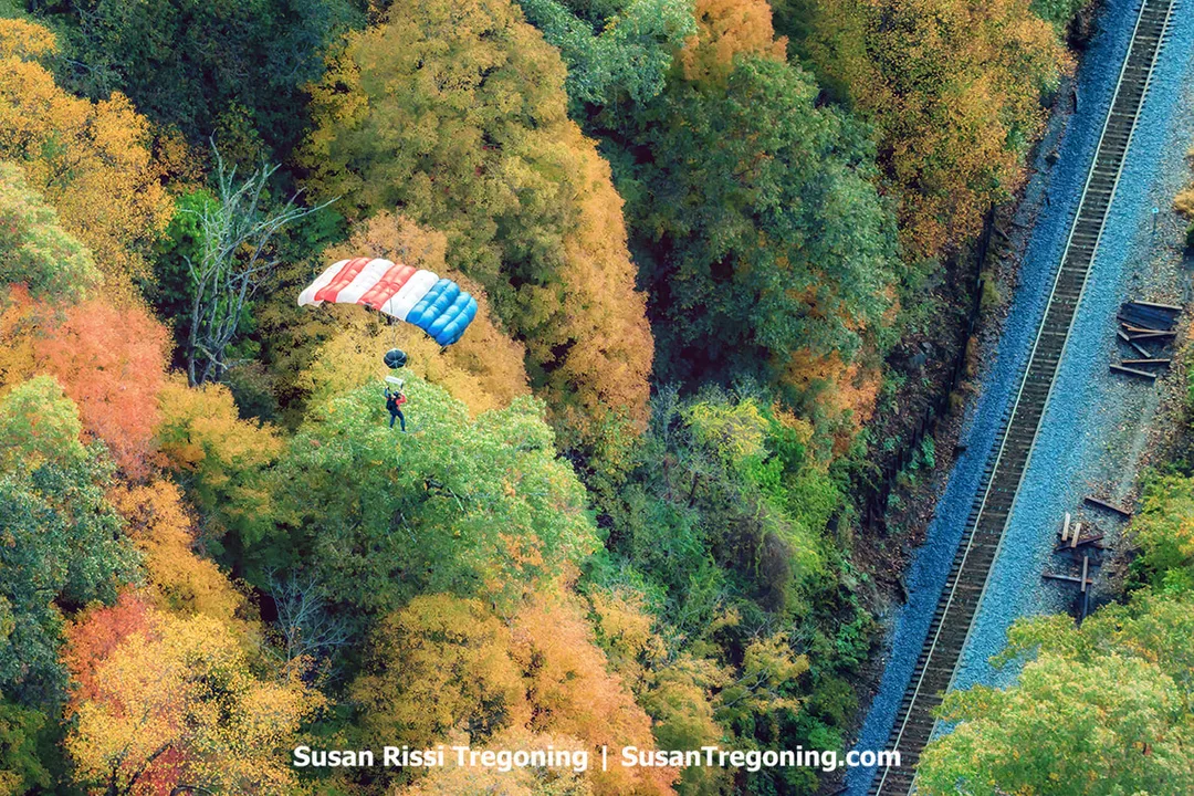 A parachutist descends beneath a red, white, and blue canopy above a forested hillside, with autumn foliage below and a rail line running through the trees to the right.