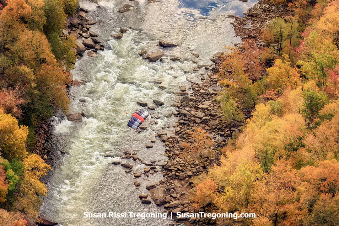 A parachute canopy with red, white, and blue panels floats above whitewater rapids in a rocky river gorge, surrounded by dense autumn foliage in shades of orange, yellow, and green.