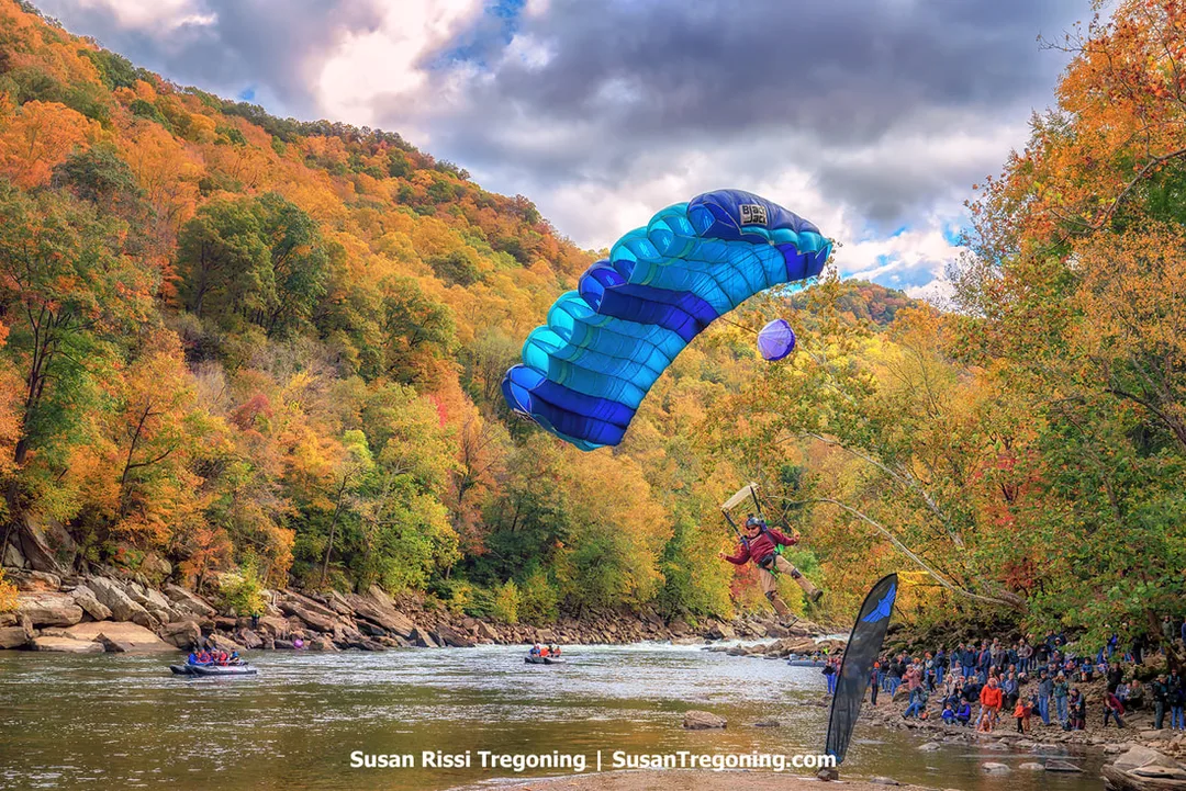 A parachutist under a bright blue and turquoise canopy descends toward a riverbank lined with spectators, with autumn‑colored hills rising behind the scene and several rafts floating on the water below.
