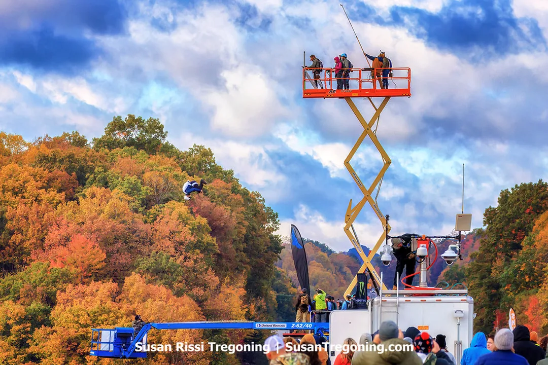 A BASE jumper mid‑somersault after launching from the New River Gorge Bridge during Bridge Day, with autumn foliage covering the surrounding hillsides and spectators gathered below near two elevated platforms used for filming and event support.