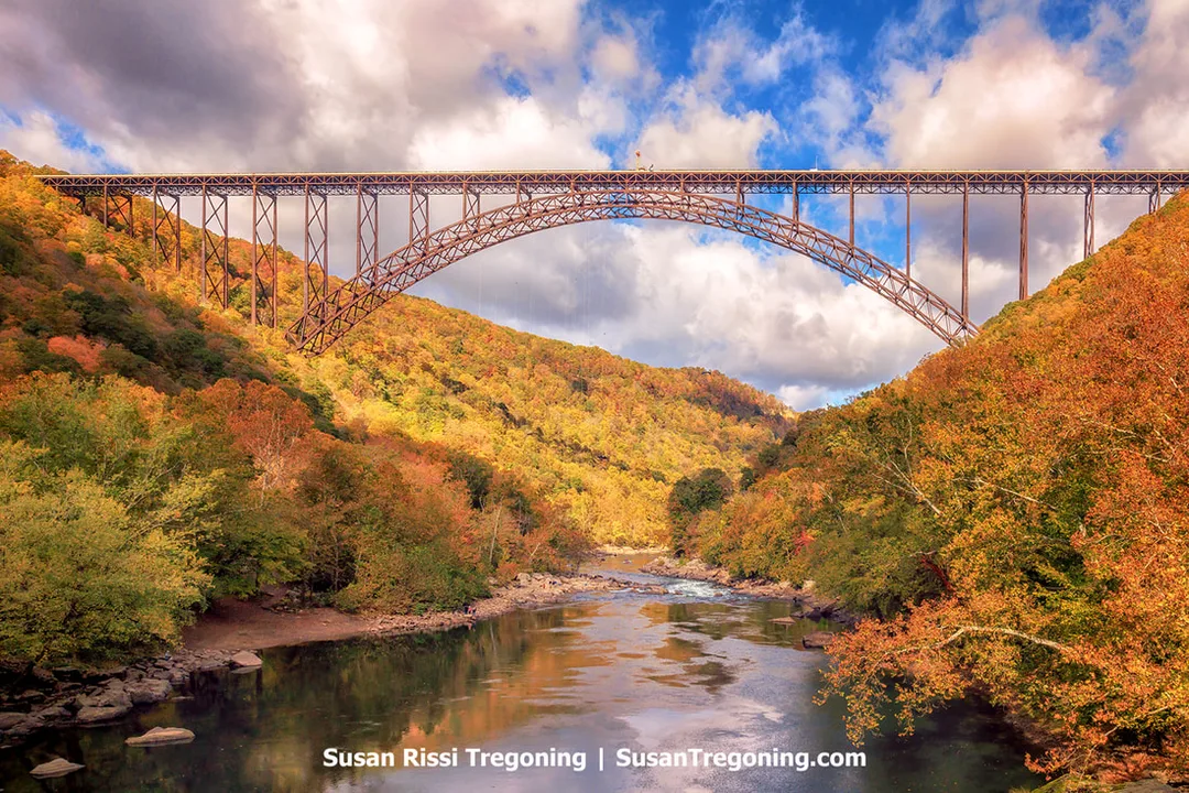 View of the New River Gorge Bridge spanning high above the river, with dense autumn foliage covering the steep hillsides and reflecting in the water below. The steel arch structure dominates the scene, captured during Bridge Day 2023.