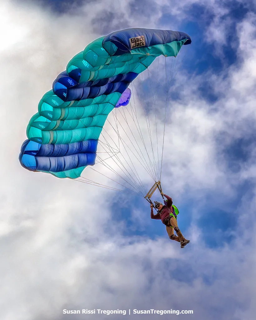 A parachutist descends beneath a blue and teal canopy against a partly cloudy sky, suspended by control lines with gear visible as the bright canopy contrasts with the shifting clouds.