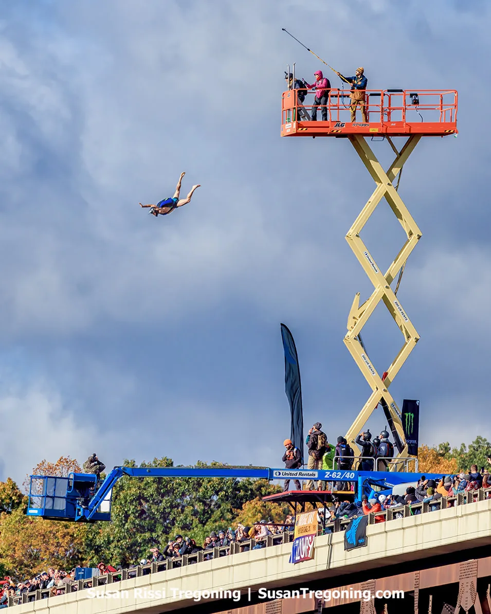   
A BASE jumper in free fall above a bridge crowded with spectators, with two elevated platforms positioned below and autumn‑colored trees filling the background under a partly cloudy sky.