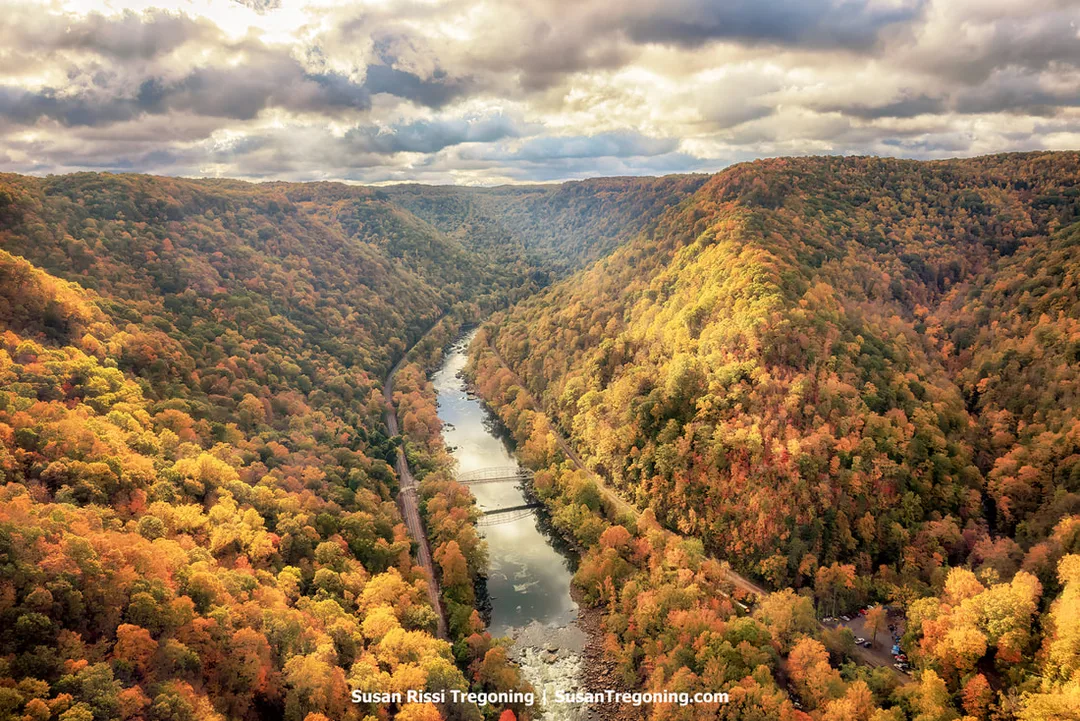 A high, aerial view of the New River Gorge in autumn, showing the steel arch of the New River Gorge Bridge crossing above a river lined with dense, multicolored foliage. The surrounding hillsides display a mix of red, orange, yellow, and green trees, with a road or rail line running along the riverbank and soft light breaking through scattered clouds.
