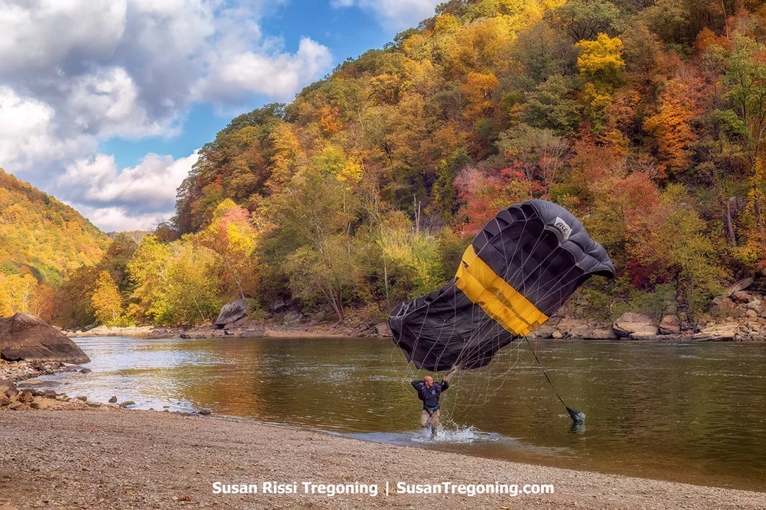 A BASE jumper runs along the riverbank beside a partially collapsed black canopy with a yellow stripe, trying to keep it out of the water as autumn foliage covers the surrounding hills.